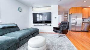 En moderne stue med sofa, skammel, fjernsyn, læderstol og et køkkenområde.