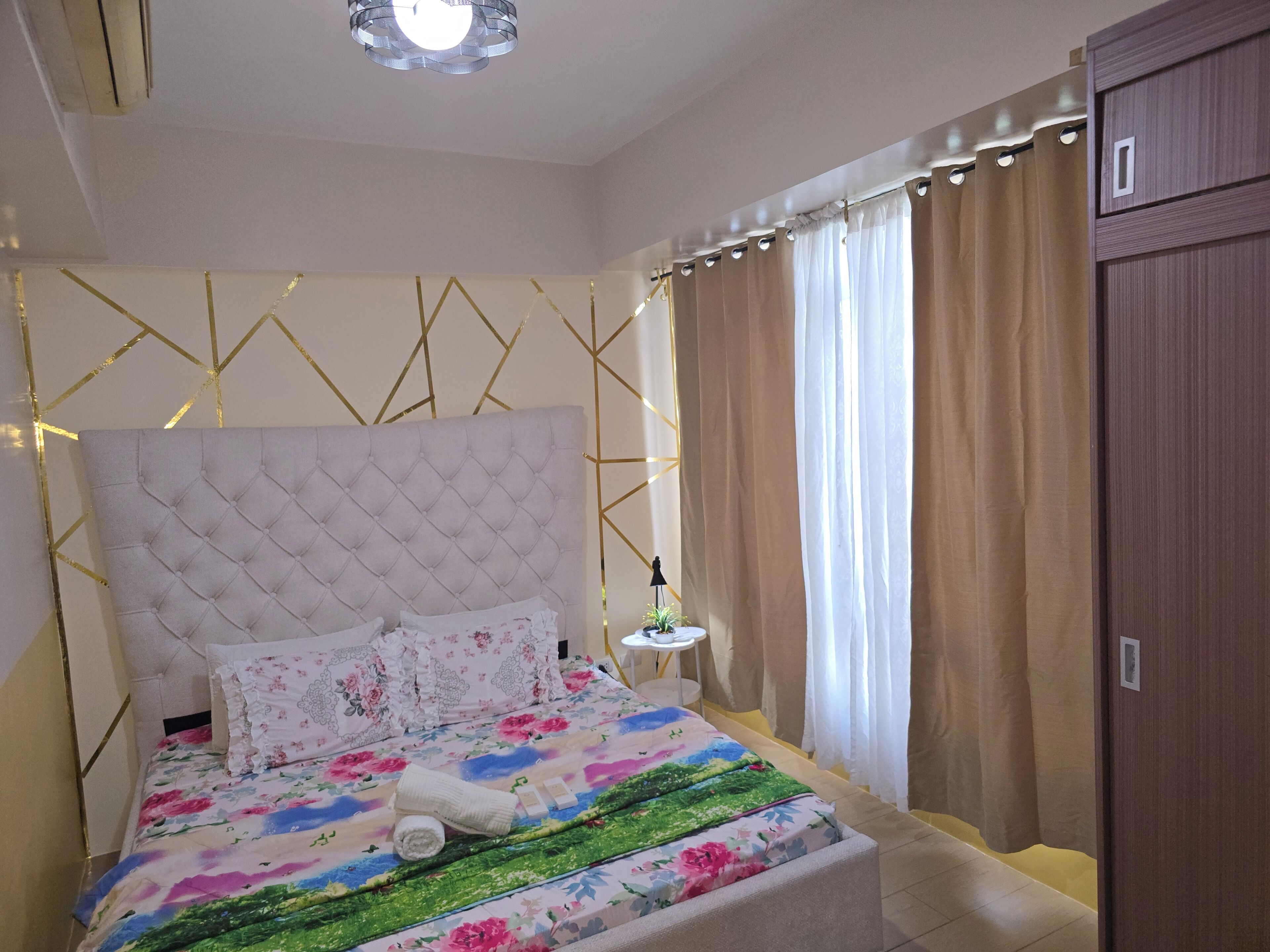 Ein ordentlich eingerichtetes Schlafzimmer mit einem gesteppten Kopfteil, floralem Bettzeug, einem Nachttisch mit Lampe und einem Kleiderschrank.