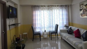 Ein Wohnzimmer mit einer Couch, zwei Sesseln, einem Fernseher, einem Deckenventilator und einem Fenster mit Vorhängen.