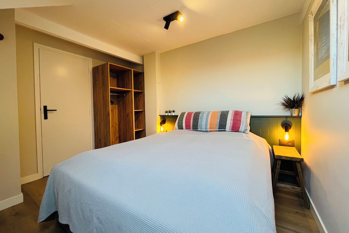 A bedroom with a bed, a wardrobe, a bedside table with a lamp, and a small stool.