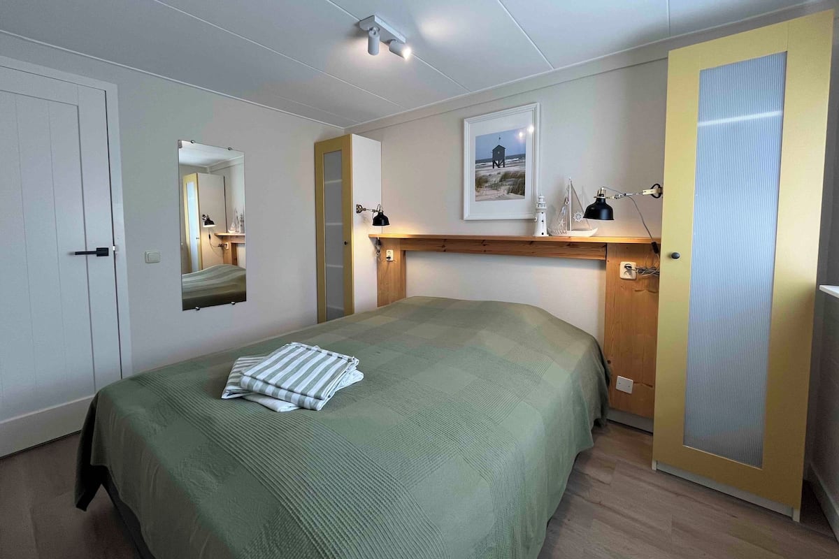 A bedroom with a bed, a bedside table, a mirror, a framed picture, and a door with a view panel.