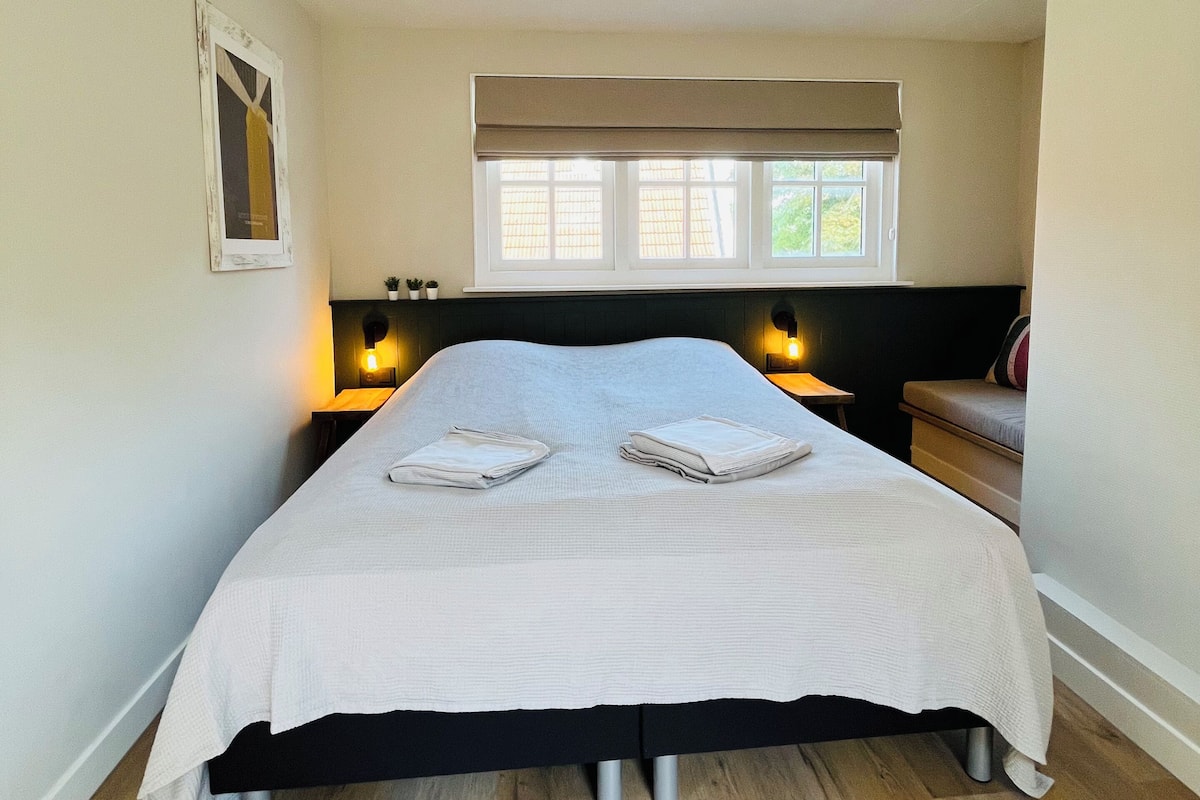 A bedroom with a bed, bedside lamps, a window with blinds, and a small potted plant.