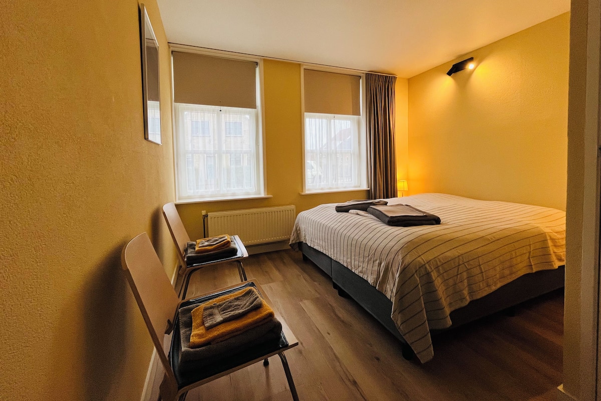 A hotel room with a bed, a chair, a radiator, and two windows.