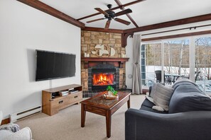 A living room with a stone fireplace, a ceiling fan, a flat-screen TV, and a sofa. - Family Condo w Summer Pool, 5 Mins to Village (Mont-Tremblant)