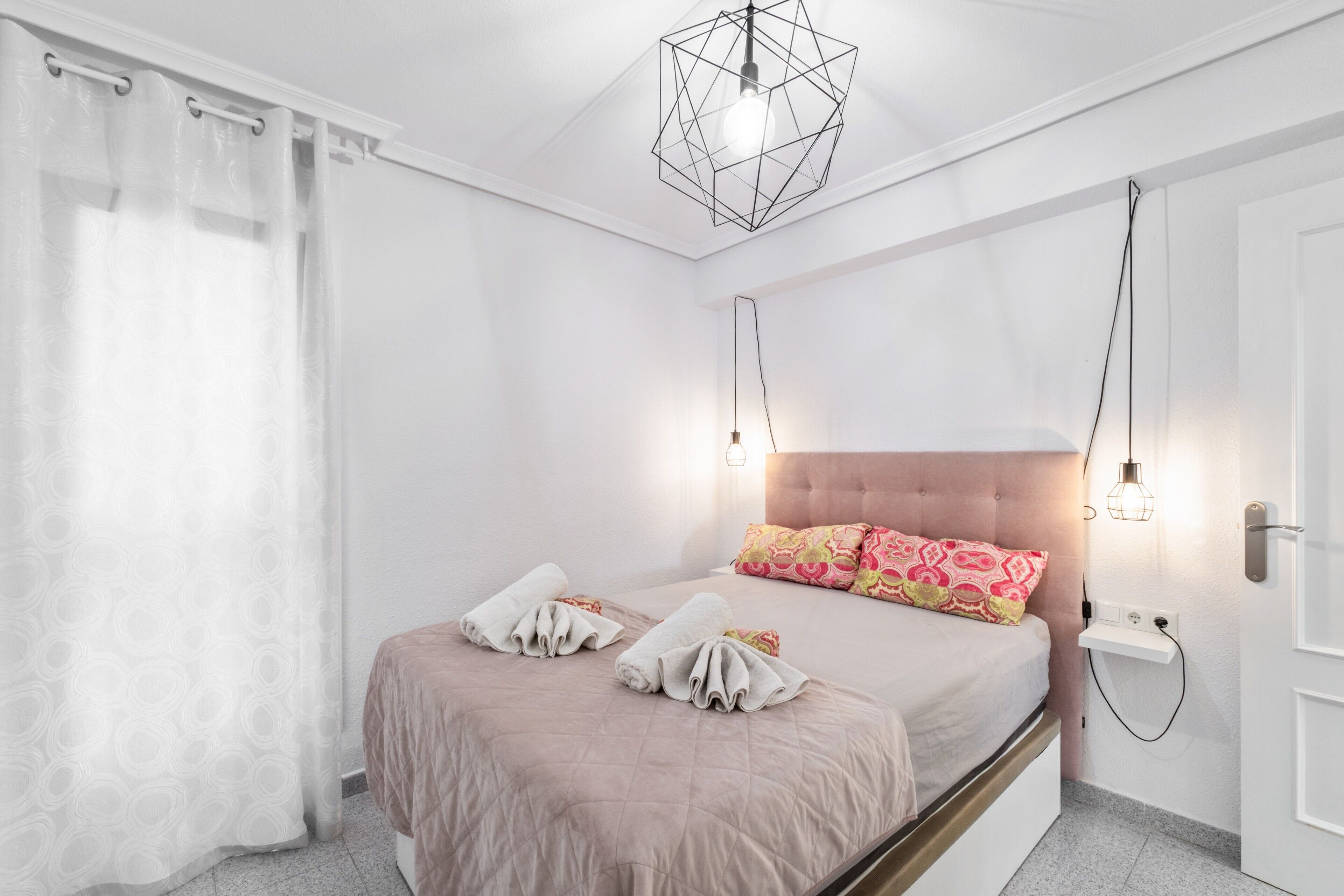 Een slaapkamer met een bed, kussens, een nachtkastje, een wandlamp en een gordijn.
