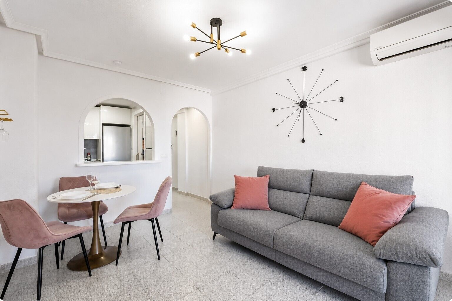 Een moderne woonkamer met een grijze bank, roze kussens, een ronde tafel met stoelen, een wandklok en een plafondlamp.