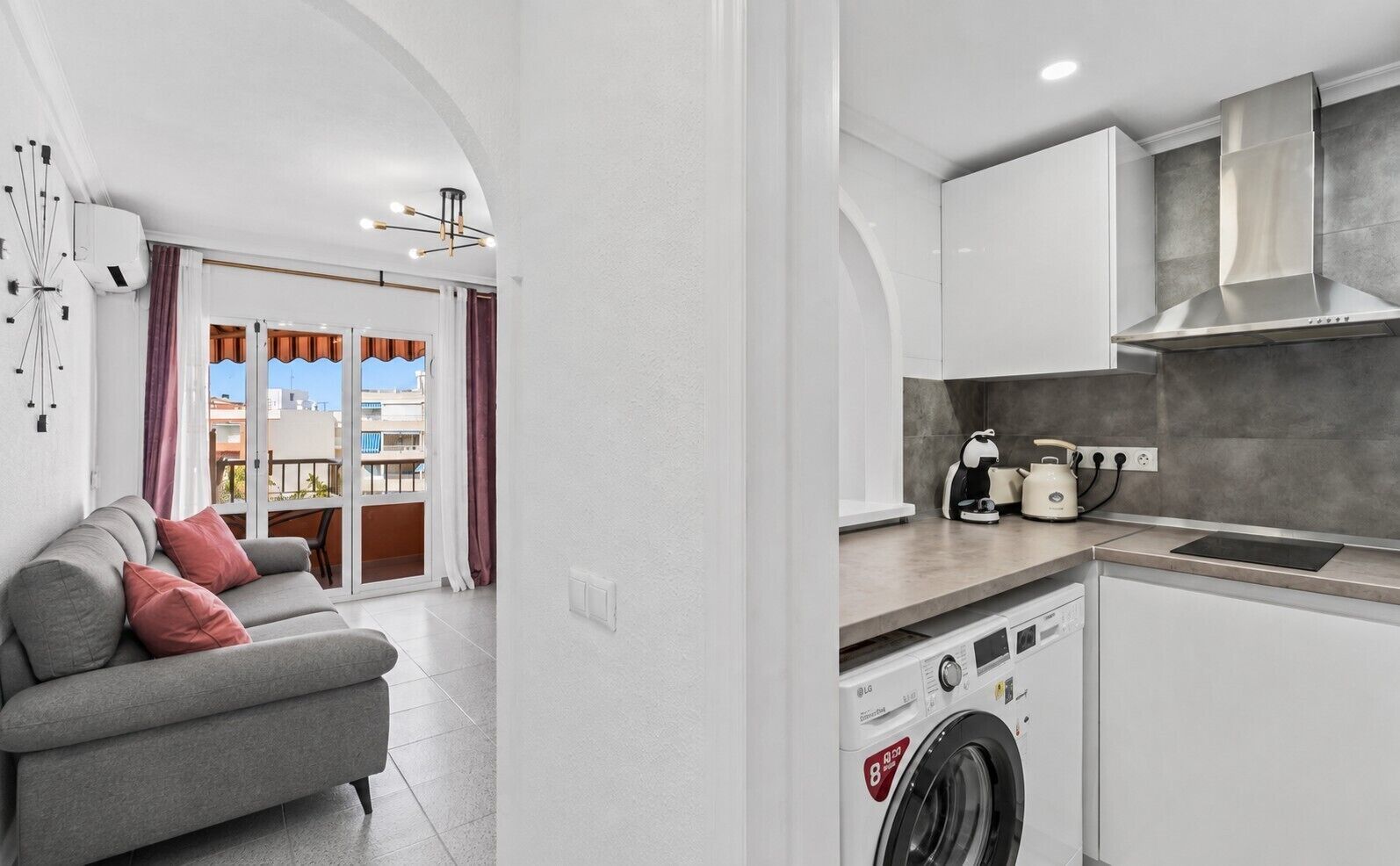 A compact living space with a grey sofa, a washing machine, and a kitchenette with a stove and kettle.