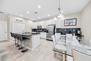 Discover this stylish kitchen with sleek white cabinetry and a spacious island ideal for casual gatherings. The elegant glass dining table invites shared meals, while the polished tile floor adds to the bright, airy atmosphere. - 4BR house with private pool in Bella Vida (Kissimmee)