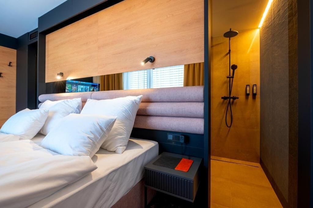 Una habitación de hotel moderna con cama, televisión integrada, ducha y una mesita de noche pequeña.