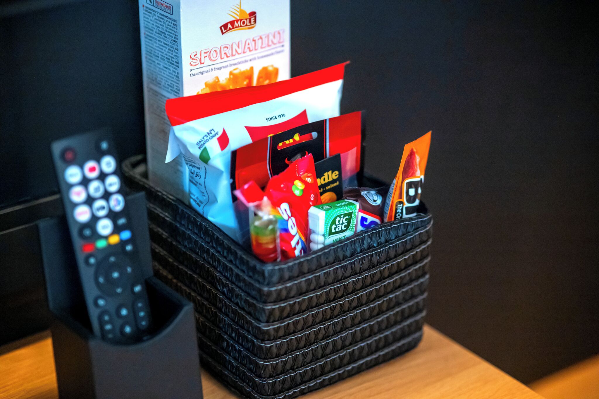 A basket containing snacks and a remote control on a wooden surface.