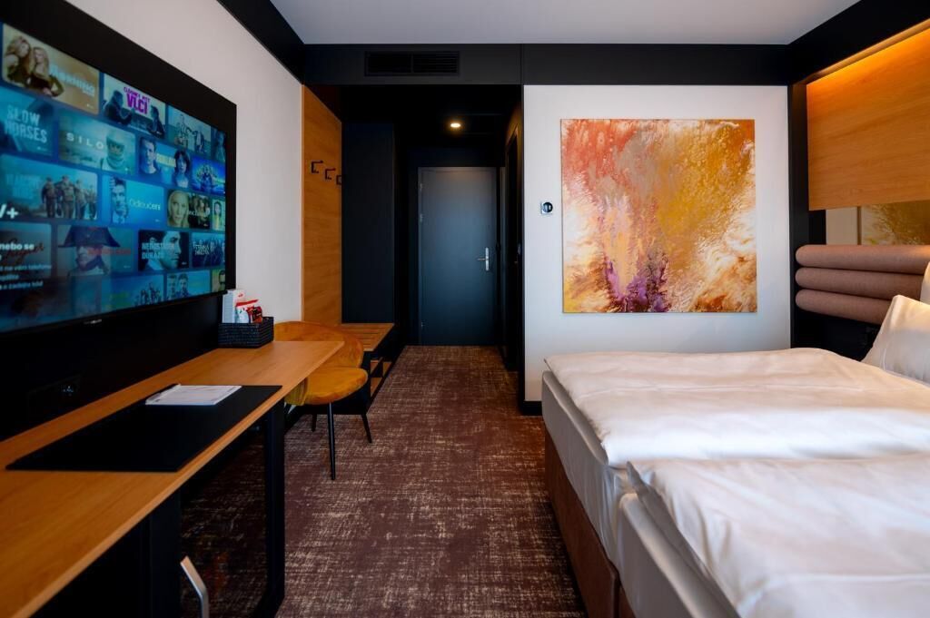A hotel room with a bed, a desk with a TV, a chair, a carpeted floor, and a large abstract painting on the wall.