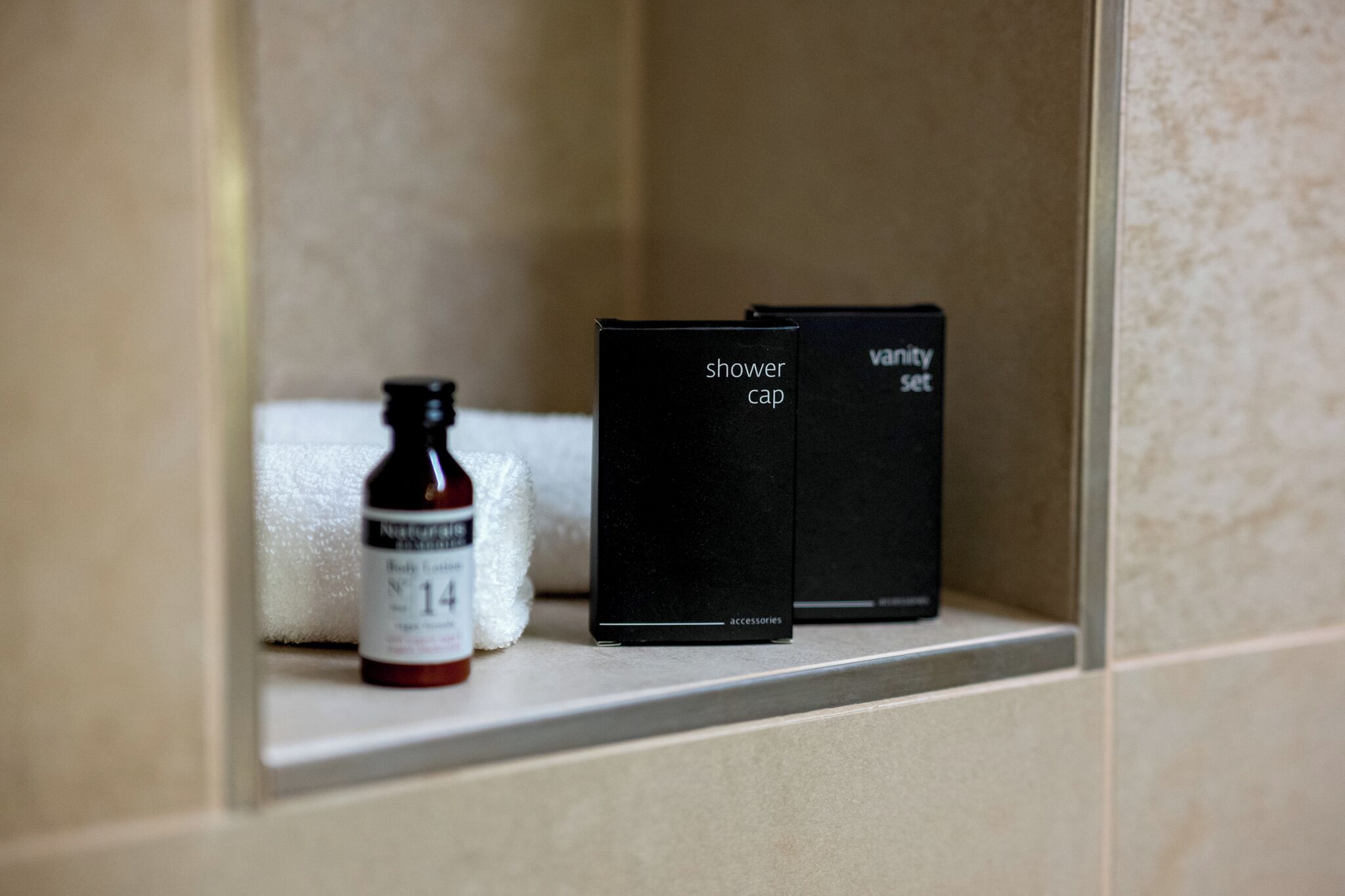 Un estante de baño con una botella de loción corporal y dos cajas negras etiquetadas como 'gorro de ducha' y 'set de tocador'.