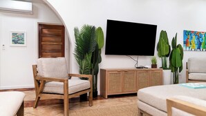 A living room with a television, a wooden cabinet, a sofa, a chair, and large green plants.