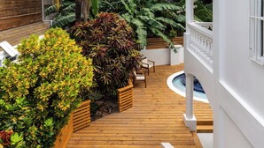 A wooden deck with a pool, surrounded by lush greenery and a white wall.