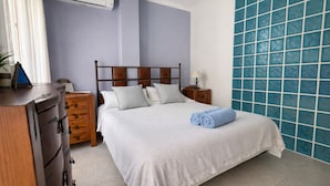 Ein ordentlich bezogenes Bett mit weißer Bettdecke und Kissen, eine hölzerne Kommode mit Schubladen, ein Nachttisch mit Lampe und eine blaue Wand mit Fliesen.