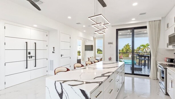 Una cocina moderna con armarios blancos, una isla de mármol y un balcón con vista a la piscina.