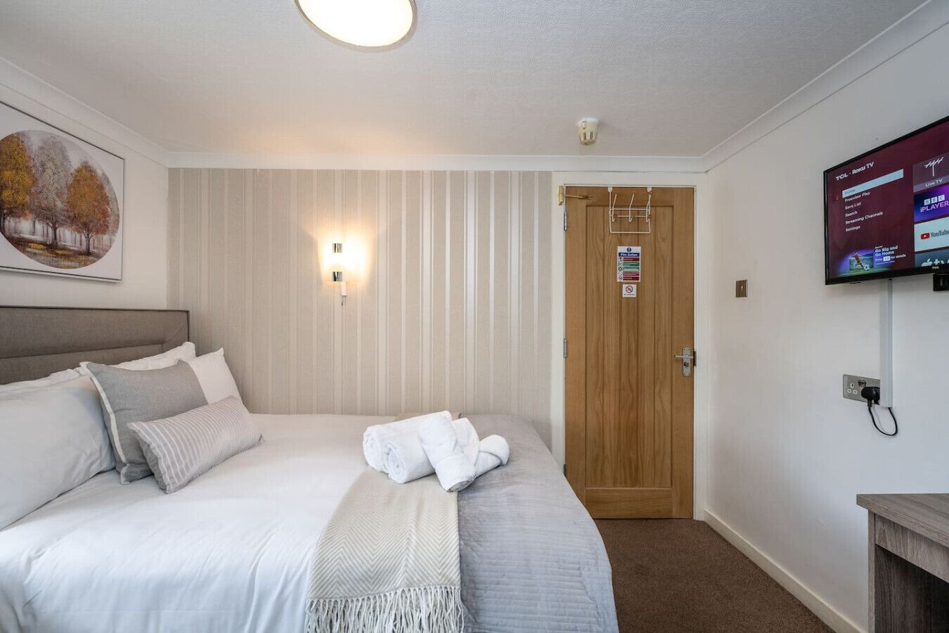 A hotel room with a bed, a wall-mounted TV, a door with a sign, and a painting on the wall.