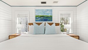 Quarto com uma cama grande, duas mesas de cabeceira, uma cabeceira de madeira, um quadro sobre a cama e uma janela com persianas.
