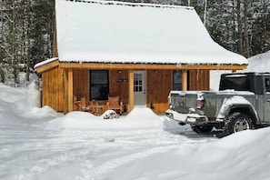 Una cabaña de madera con techo nevado, un porche con sillas y un vehículo estacionado.