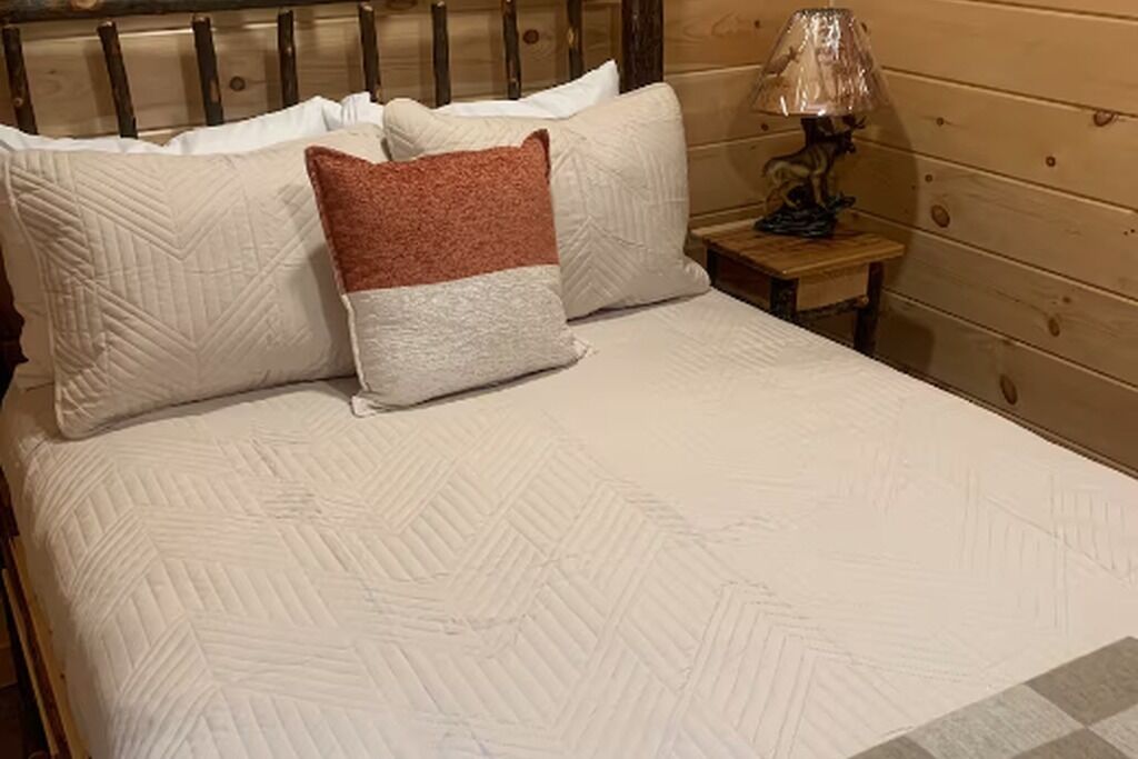 Una cama con cubrecolcha blanco acolchado, dos almohadas (una con detalle marrón) y una lámpara de noche sobre una mesita de madera.