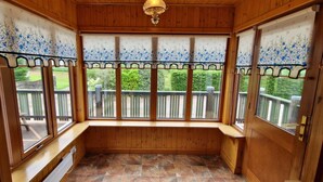 A wooden-paneled room with a bench, large windows with decorative curtains, and a view of a wooden deck and greenery outside.