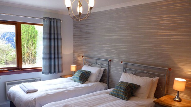 A hotel room with two beds, a window with curtains, a wall-mounted heater, and a chandelier.