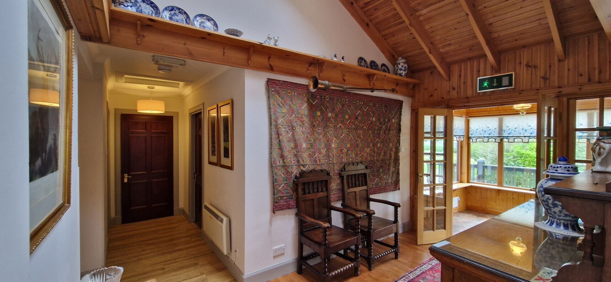A room with wooden beams, a patterned tapestry, two wooden chairs, a table with a vase, and a view through a window.