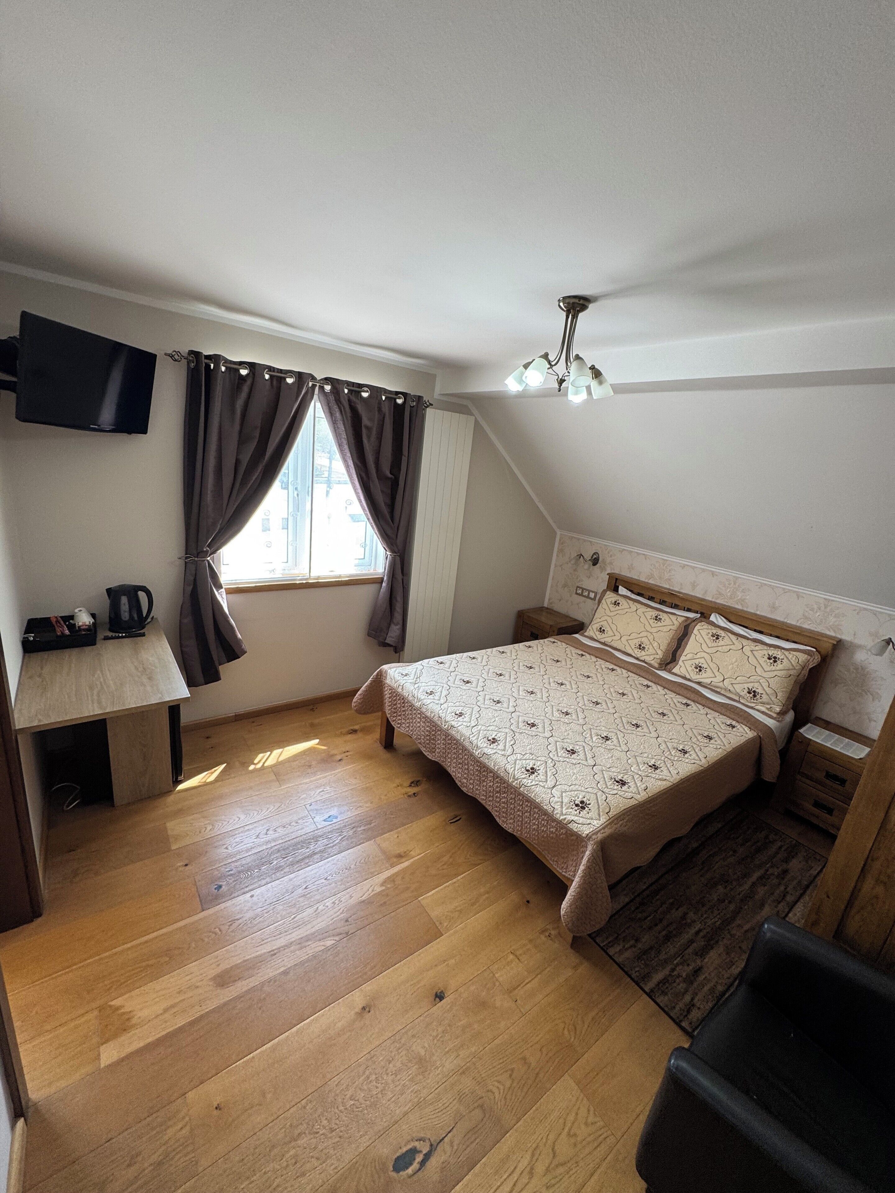 Une chambre à coucher comprenant un lit, un bureau, une chaise, un téléviseur fixé au mur et une fenêtre avec des rideaux.