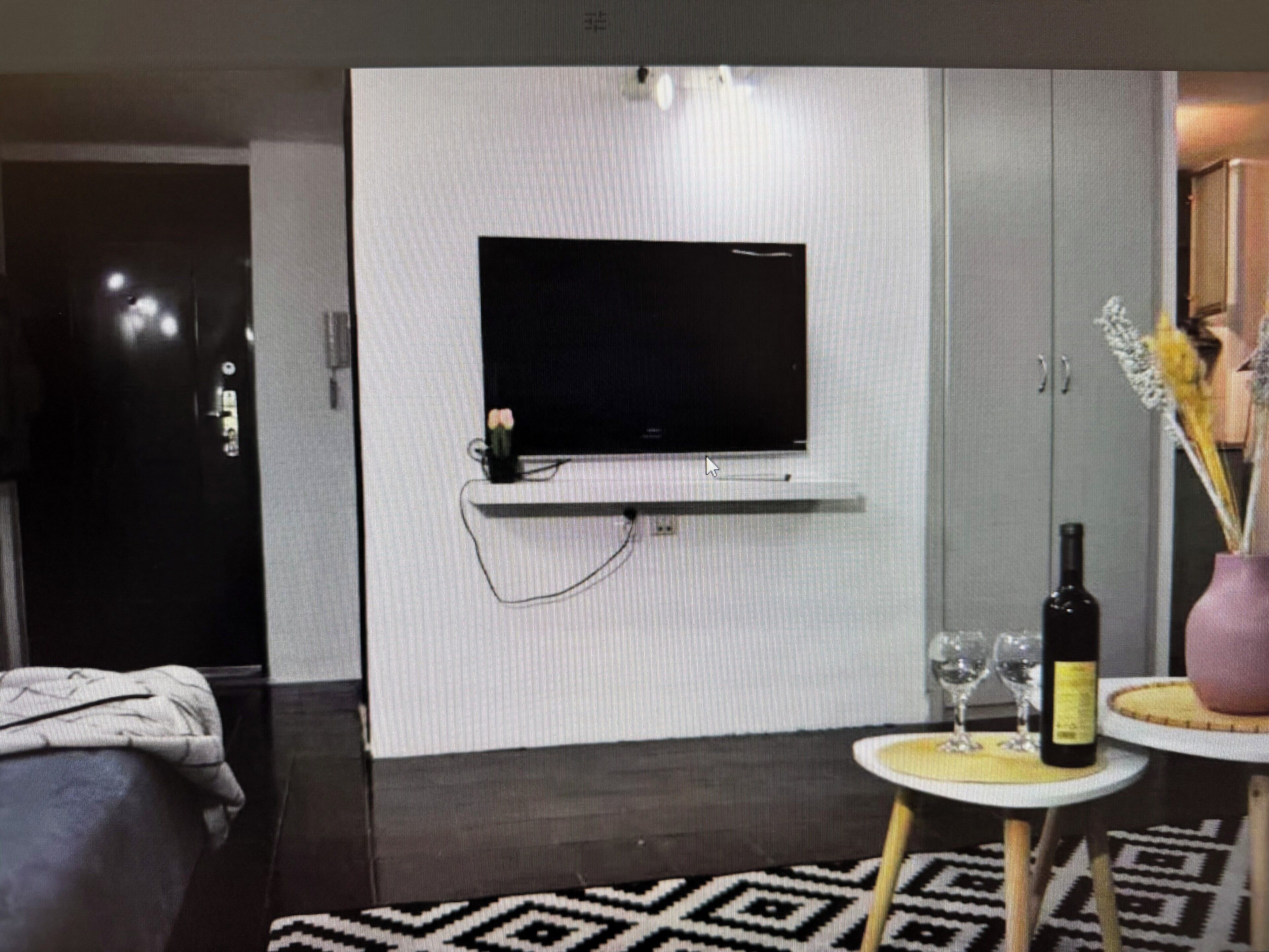 Una habitación moderna con un televisor de pantalla plana en una pared blanca, una mesita redonda con una botella y vasos, y un tapete estampado en el suelo.