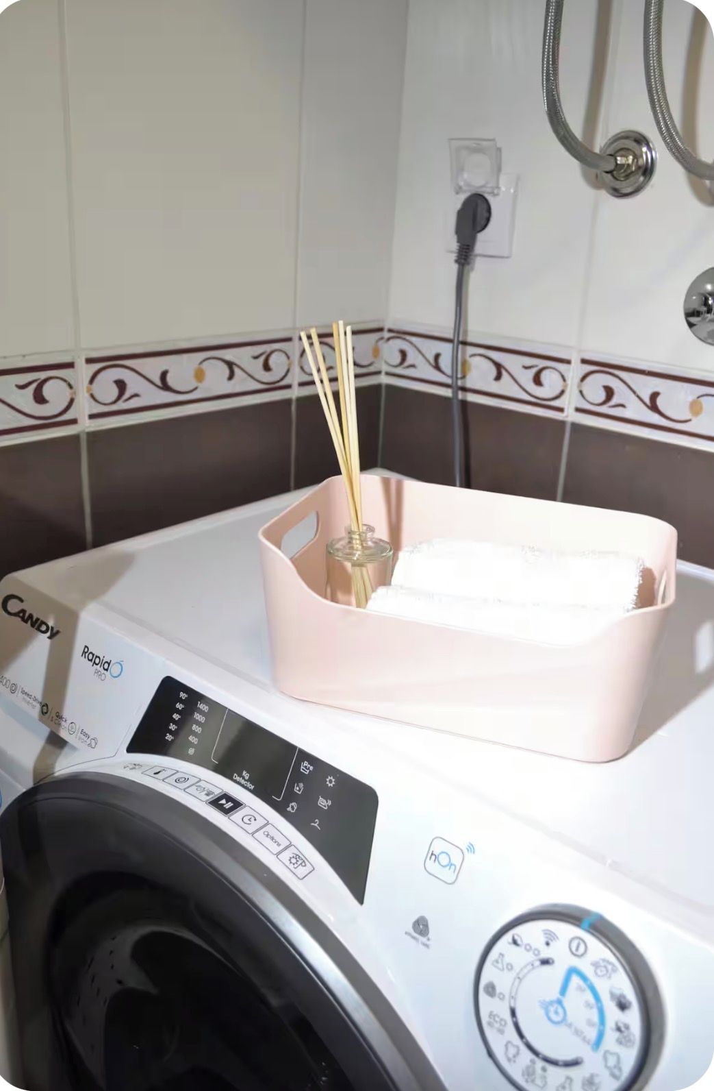 Une machine à laver blanche dotée d’un panneau de commande, un panier rose contenant un récipient en verre, et un pare-éclaboussures décoratif en carreaux.