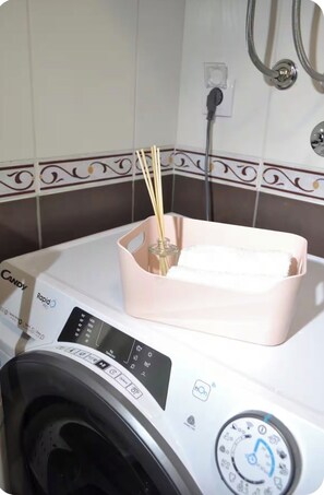 A white washing machine with a control panel, a pink basket with a glass container, and a decorative tile backsplash.