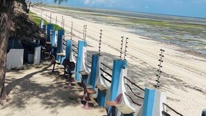 Een zitgedeelte aan het strand met stoelen en tafels, een rieten dakconstructie en uitzicht op de oceaan.