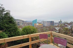 A wooden deck with two chairs and a table, overlooking a cityscape with buildings and a distant hill. - Contractor Stay • Deck • TVs in Every Room (Swansea)