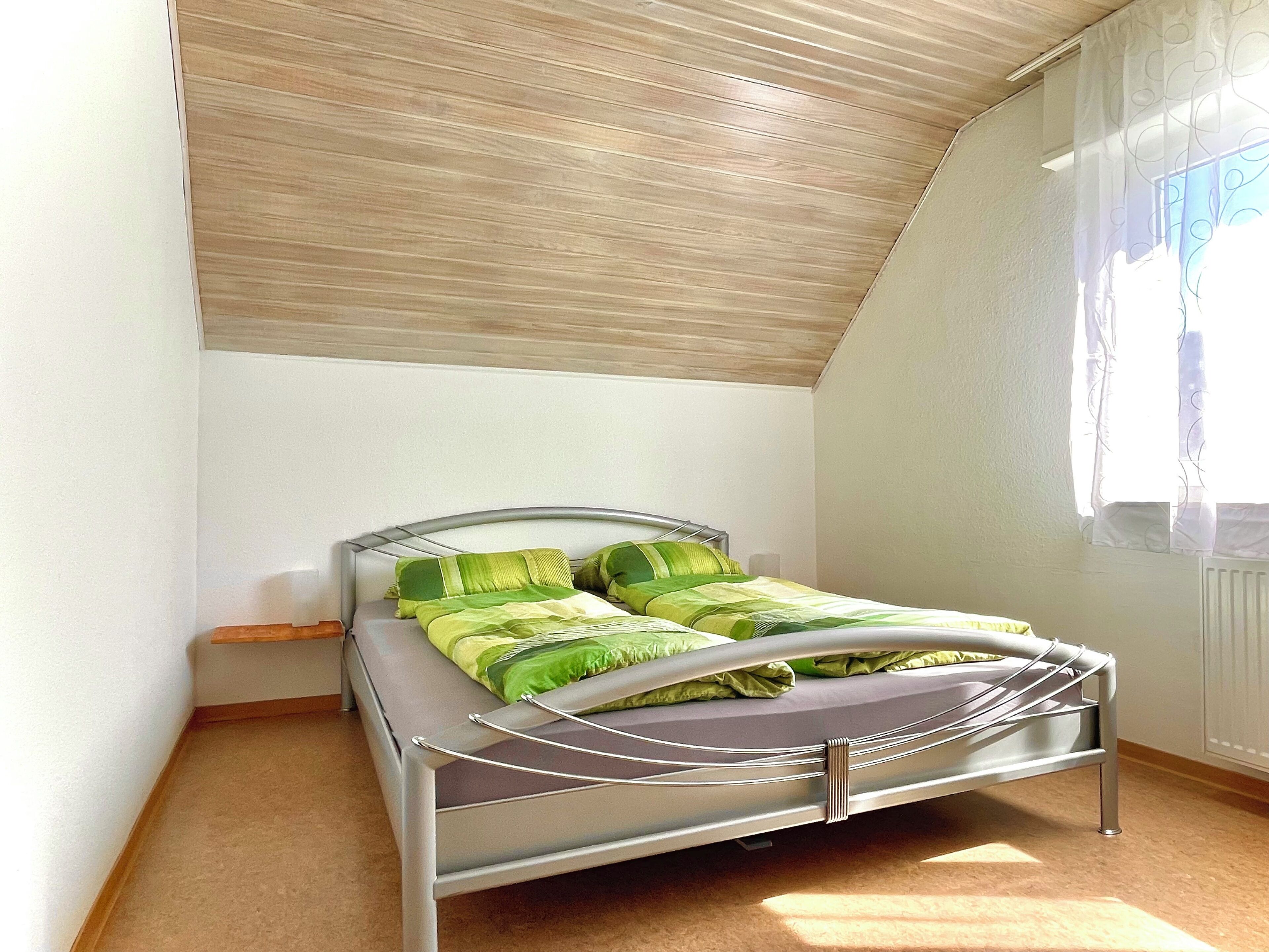 Una cama individual con estructura metálica y cabecera, una ventana con cortinas translúcidas y un techo inclinado.