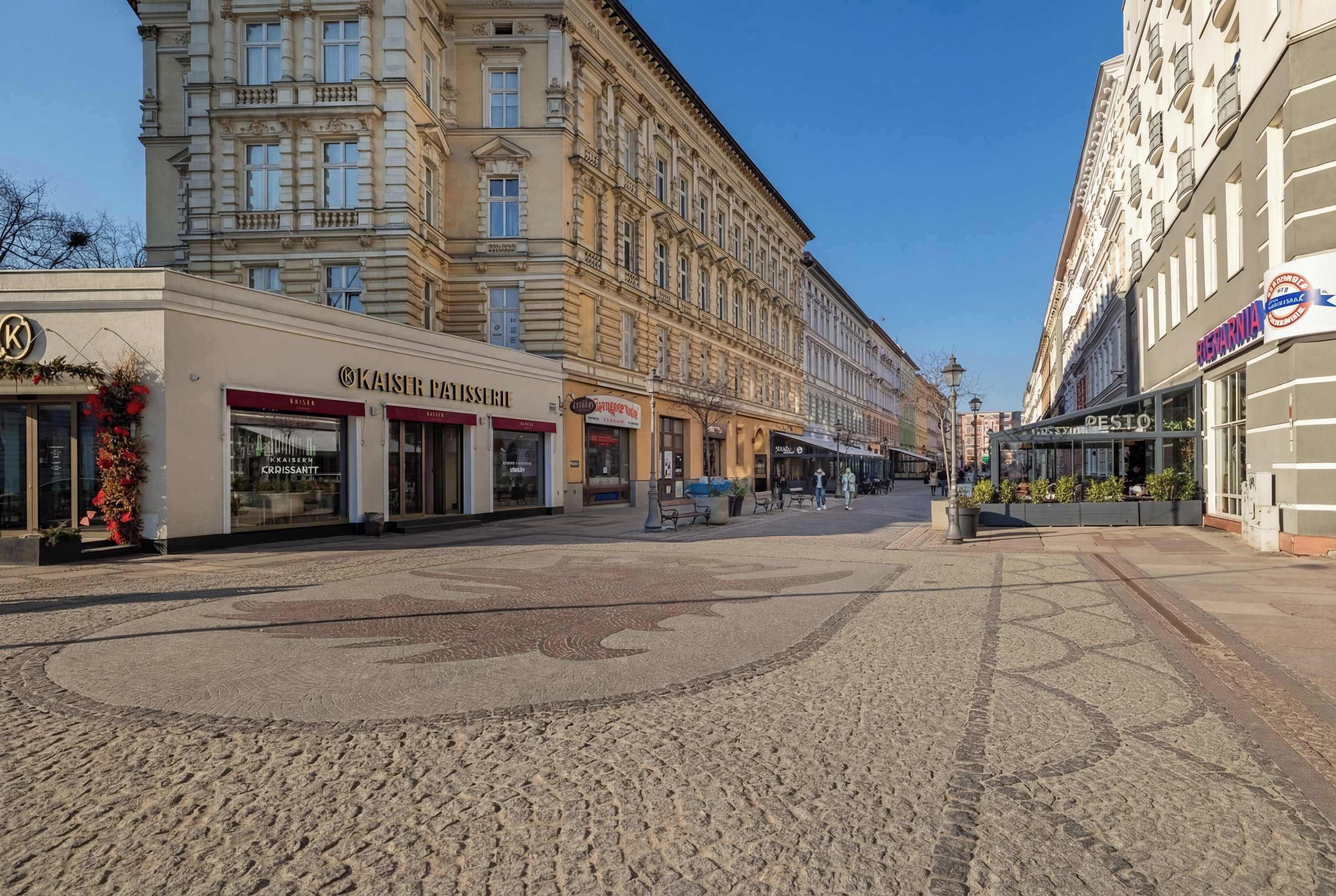 Eine Kopfsteinpflasterstraße mit Gebäuden an beiden Seiten, darunter ein Restaurant namens 'Kaiser Patisserie' und ein Gebäude mit dem Schriftzug 'RESTAURANT'.