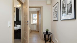 Um corredor com porta de madeira que dá para um banheiro, uma mesinha com uma planta e duas fotos emolduradas na parede.