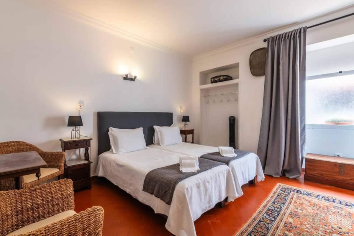 A hotel room with two beds, a small table, wicker chairs, a rug, a wardrobe, and a window with curtains.