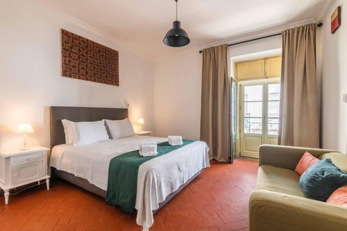 A hotel room with a bed, bedside lamps, a green bedspread, a sofa, and a view of the outside.
