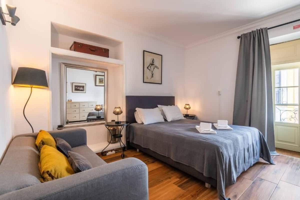 A hotel room with a bed, a sofa, a desk, a lamp, a mirror, and a window with curtains.