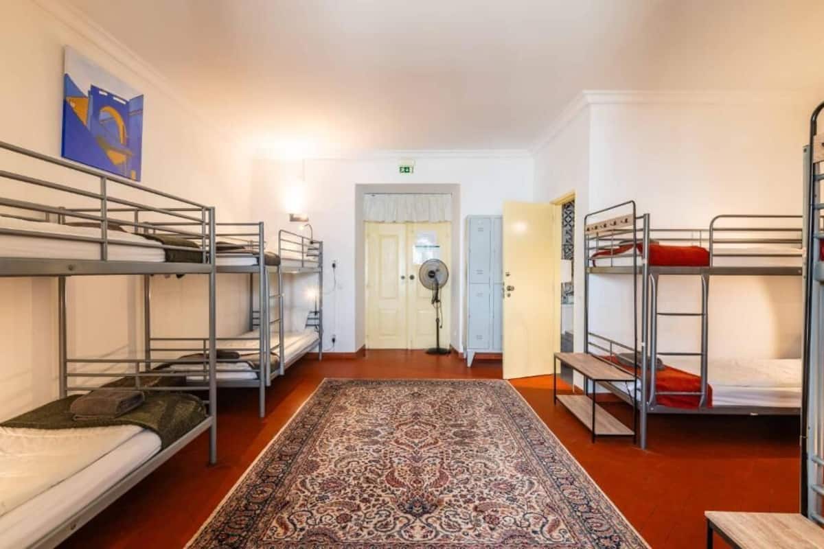 A dormitory room with bunk beds, a patterned rug, a fan, and a door with an exit sign.