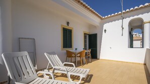 A sunlit patio with white lounge chairs, a small table, and chairs, a tiled floor, and a white building with a tiled roof and a window with shutters. - U 01 - Bedroom 1 in Casa Quinta Estação (Lagos)
