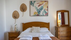 A neatly made bed with white linens, a wooden headboard, a bedside table with a lamp, a mirror, and a wall art piece.