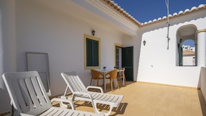 A sunlit patio with white lounge chairs, a small table, and chairs, a tiled floor, and a white building with a tiled roof and a window with shutters.