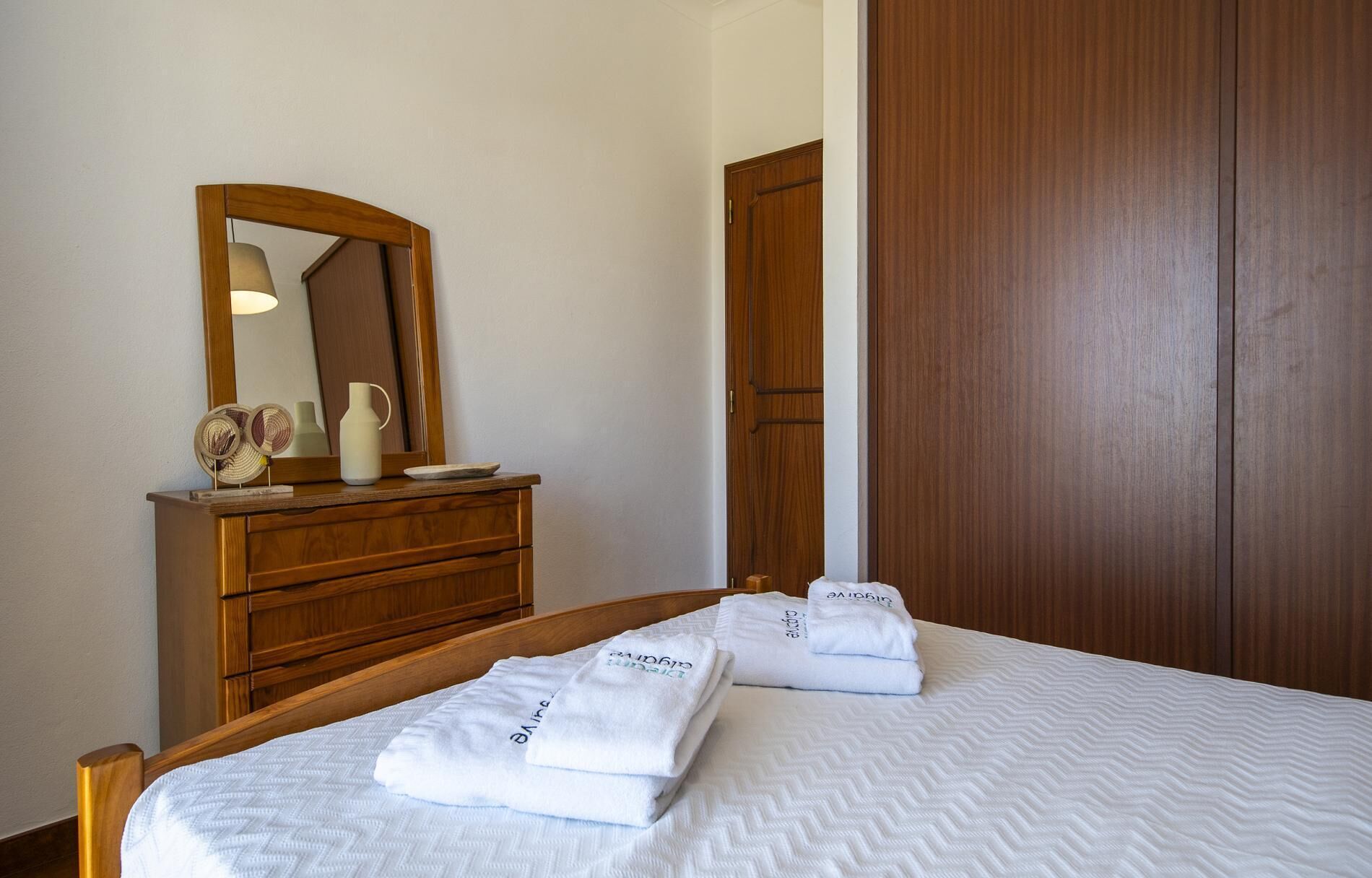 Une chambre d’hôtel avec une commode en bois, un miroir, un lit avec une literie blanche et des serviettes avec du texte imprimé.