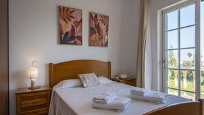 A neatly made bed with folded towels, a bedside lamp, and a window with a view of greenery.