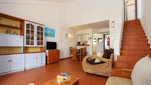Un salon comprenant un canapé, un fauteuil en osier, un téléviseur et un coin repas avec des chaises et une table.