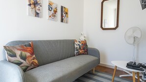A grey sofa with floral cushions, a white wall with three artworks, a wooden-framed mirror, and a standing fan. - S 01 - 1 Bed Apartment in Quinta Silvestre (Lagos)