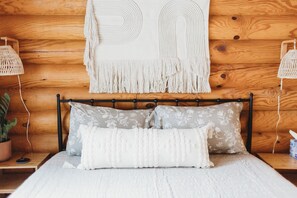 Ein Bett mit weißer, strukturierter Bettdecke, zwei Kissen mit Blumenmuster und einem Wandbehang aus Makramee.