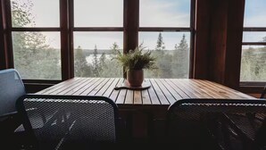 Une table en bois, une plante en pot, deux chaises en métal ajouré et de grandes fenêtres donnant sur des arbres et le ciel.