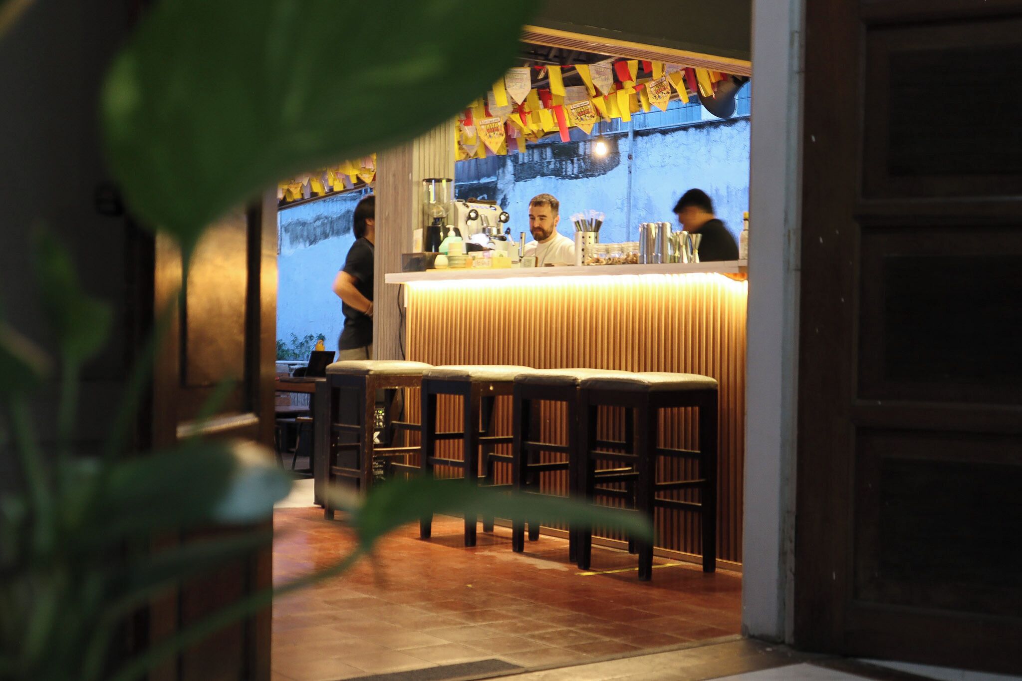 Uma área de bar com bancos e balcão, uma pessoa em pé atrás do balcão, e outra pessoa ao fundo.
